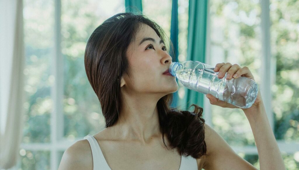 Asian woman in a sports bra drinking from a water bottle indoors.