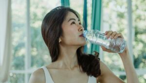 Asian woman in a sports bra drinking from a water bottle indoors.